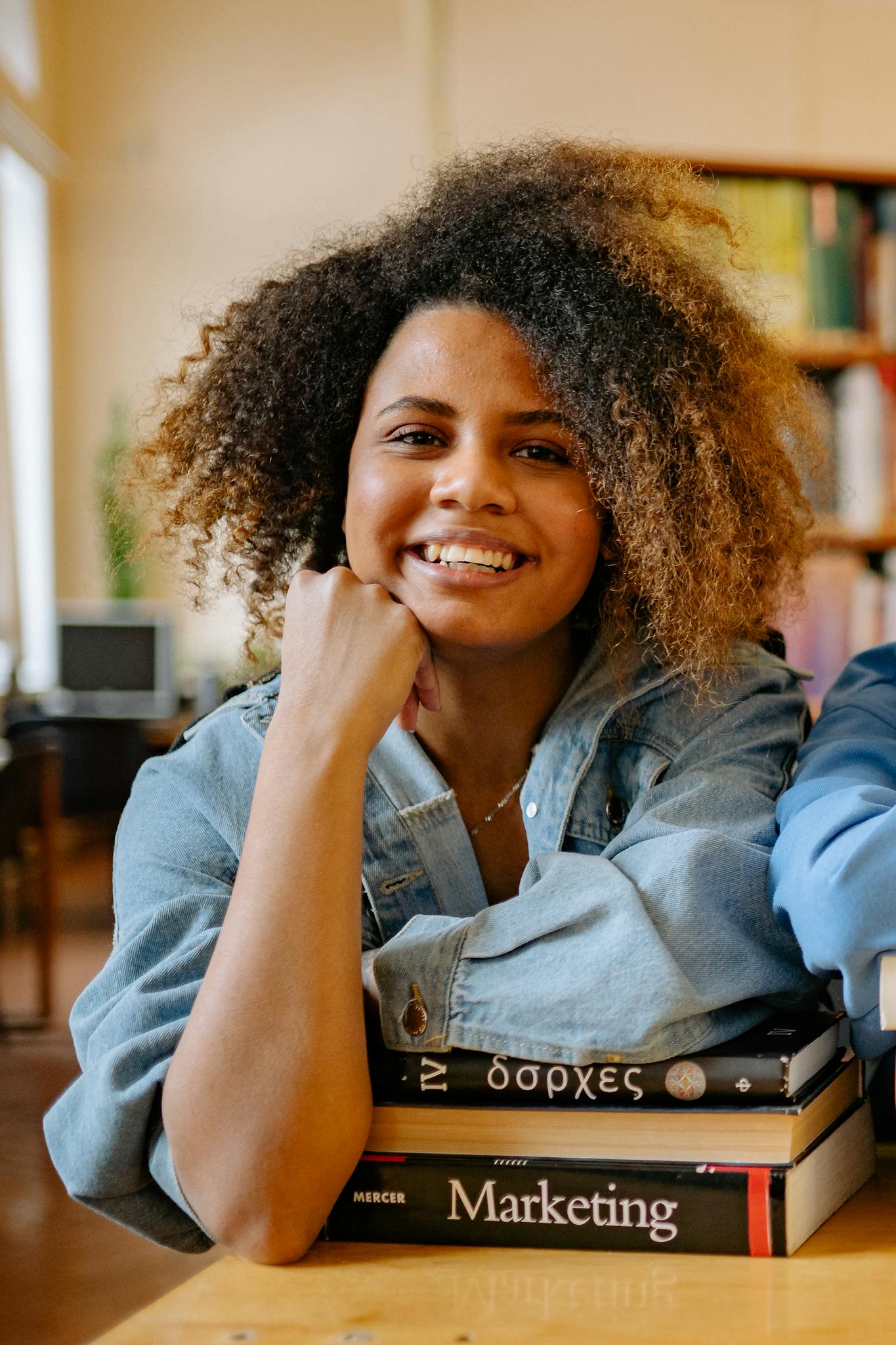 African American student smiling indoors with books, wearing a denim jacket.